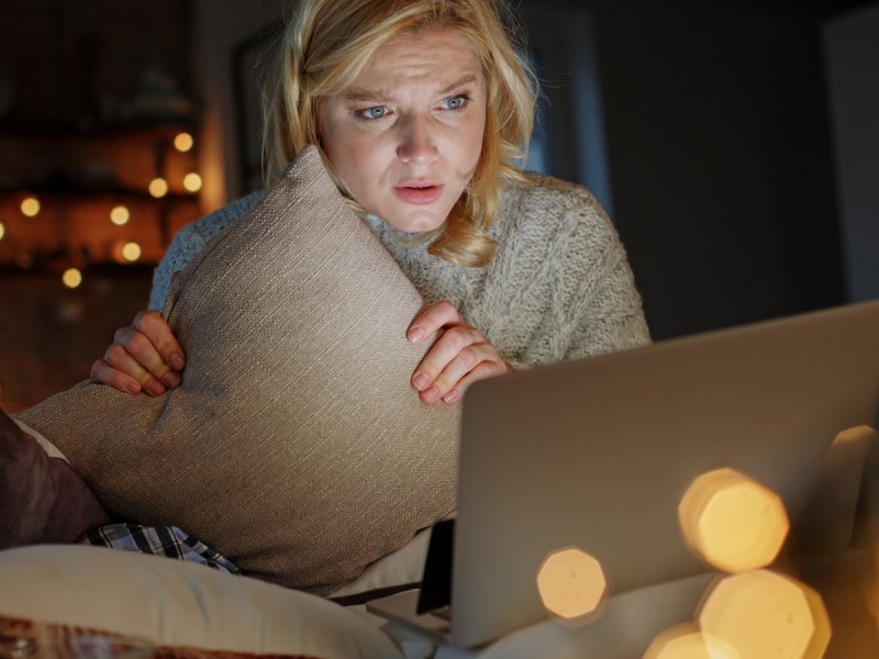 Frau sitzt mit einem Kissen angstvoll vor ihrem Laptop.