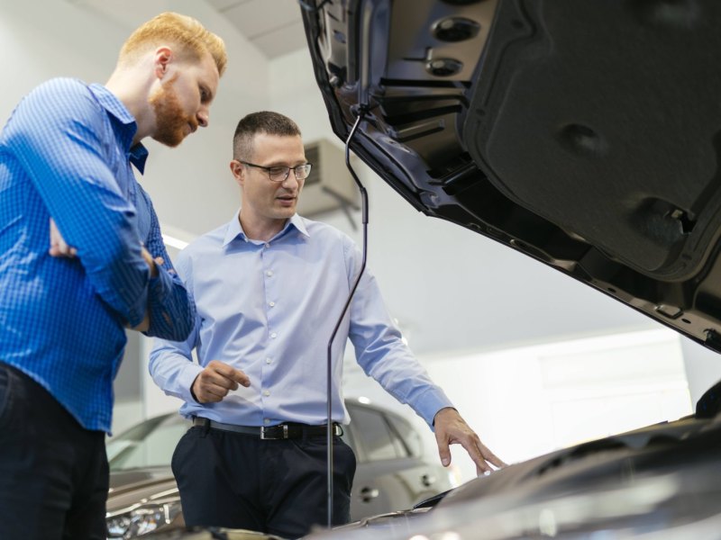 Verkäufer und Kunde in einem Autohaus schauen sich den Motor eines Wagens an.