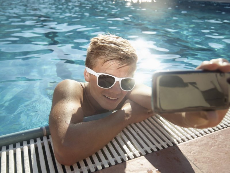 Ein junger Mann mit Sonnenbrille lehnt am Schwimmbeckenrand und macht ein Selfie.