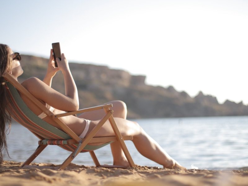 Eine Frau liegt am Strand in einer Liege und schaut auf ihr Smartphone.