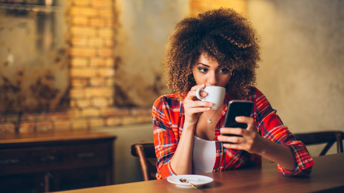 Frau mit Handy trinkt aus einer Tasse.