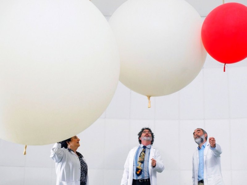 Menschen in Laborkitteln stehen unter drei Luftballons