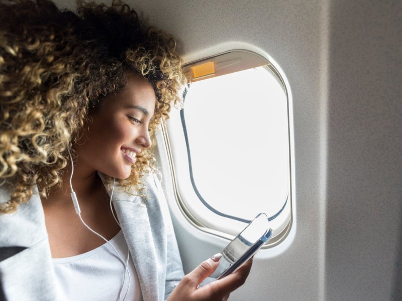 Frau sitzt mit Handy und Kopfhörern am Fenster im Flugzeug.