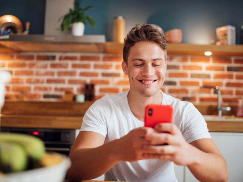 Junger blonder Mann mit Handy in der Küche.