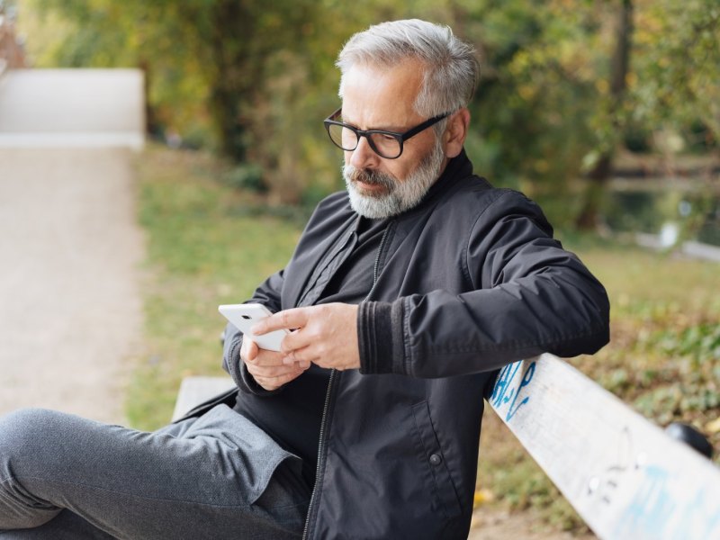 Mann sitzt mit iPhone auf Parkbank.