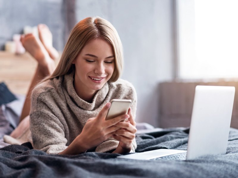 Eine Frau liegt vor ihrem Laptop und schaut aufs Handy.