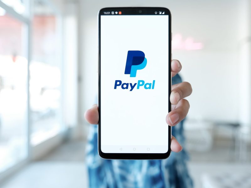 Eine Person hält ein Handy. Auf dem Display wird das PayPal-Logo angezeigt.