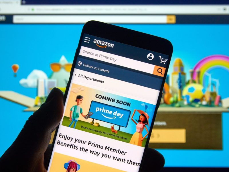 Auf einem Handy-Display wird eine Ankündigung für den Amazon Prime Day angezeigt. Im Hintergrund ist ein Computerbildschirm zu sehen, auf dem Amazon geöffnet ist.