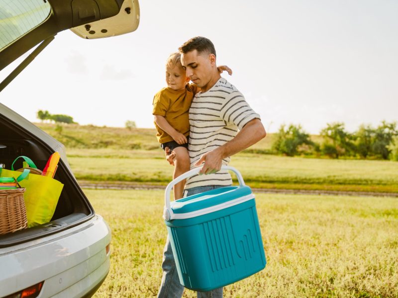 Mann trägt Kind und eine Auto-Kühlbox zu seinem Kofferraum.