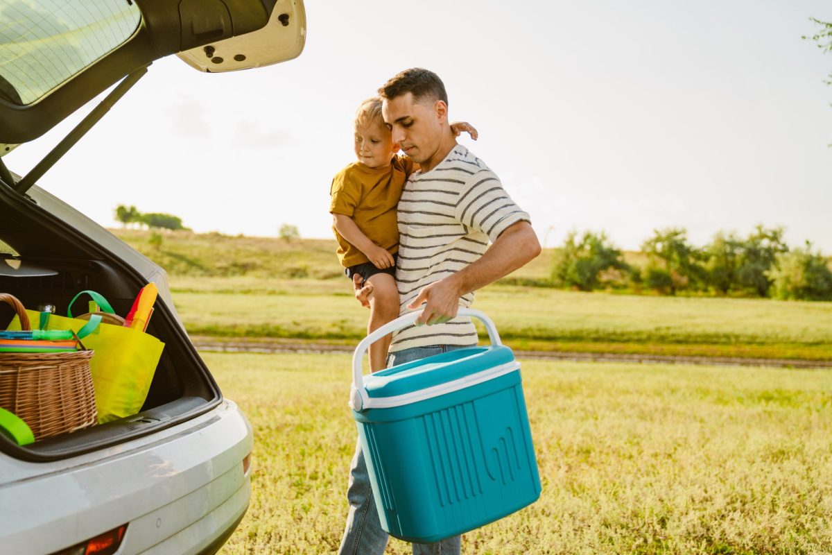 Mann trägt Kind und eine Auto-Kühlbox zu seinem Kofferraum.