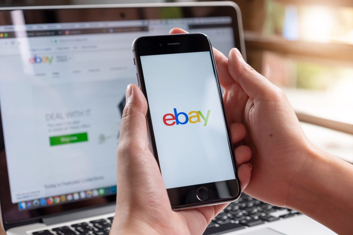Person hält Handy mit eBay-Logo auf dem Display. Im Hintergrund ist die eBay-Website auf einem Laptop offen.