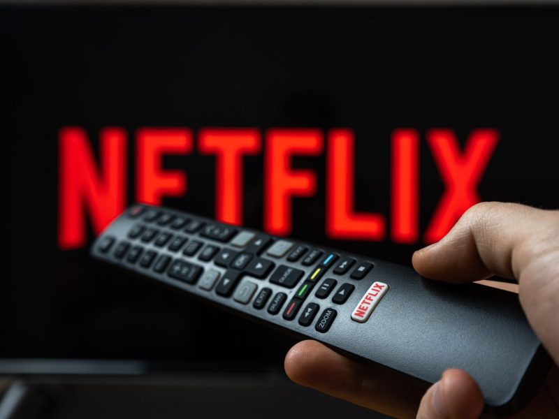 Eine Person hält eine Fernbedienung. Im Hintergrund wird auf einem Fernseher das Netflix-Logo angezeigt.