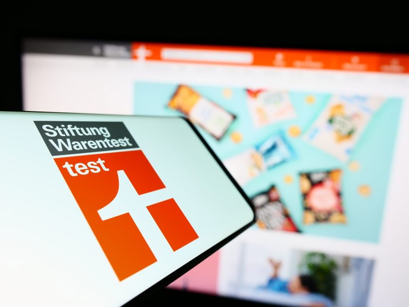 Stiftung-Warentest-Logo auf Smartphonebildschirm, im Hintergrund ein Bildschirm mit Stiftung Warentest Website