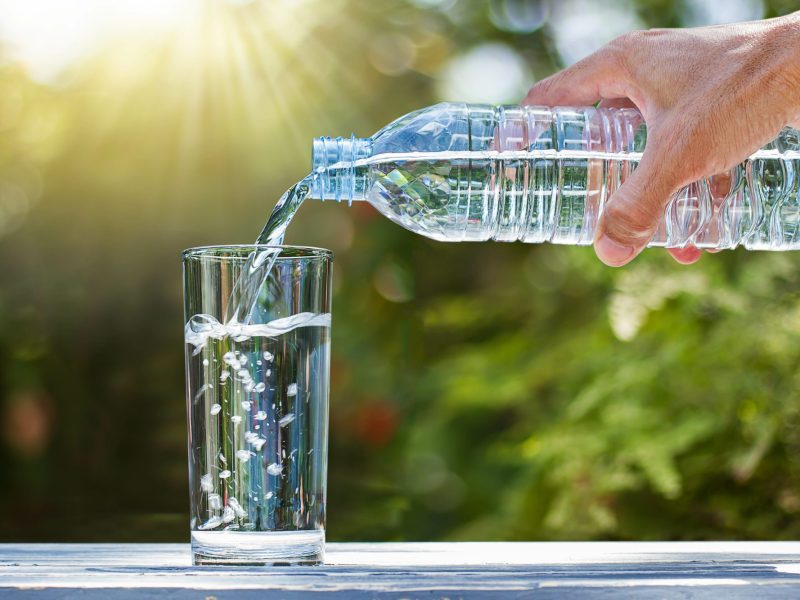 Eine Person gießt Wasser aus einer Plastikflasche in ein Glas. Im Hintergrund ist Natur und Sonnenschein zu sehen.