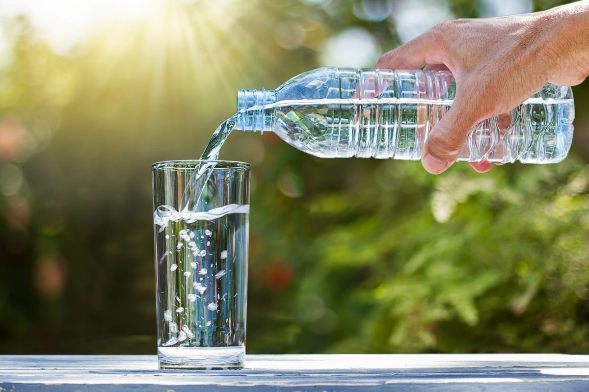 Eine Person gießt Wasser aus einer Plastikflasche in ein Glas. Im Hintergrund ist Natur und Sonnenschein zu sehen.