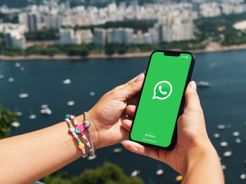 Eine Person hält ein Handy in der Hand, auf dessen Display das WhatsApp-Logo angezeigt wird. Im Hintergrund ist das Meer und die Skyline einer Stad zu sehen.