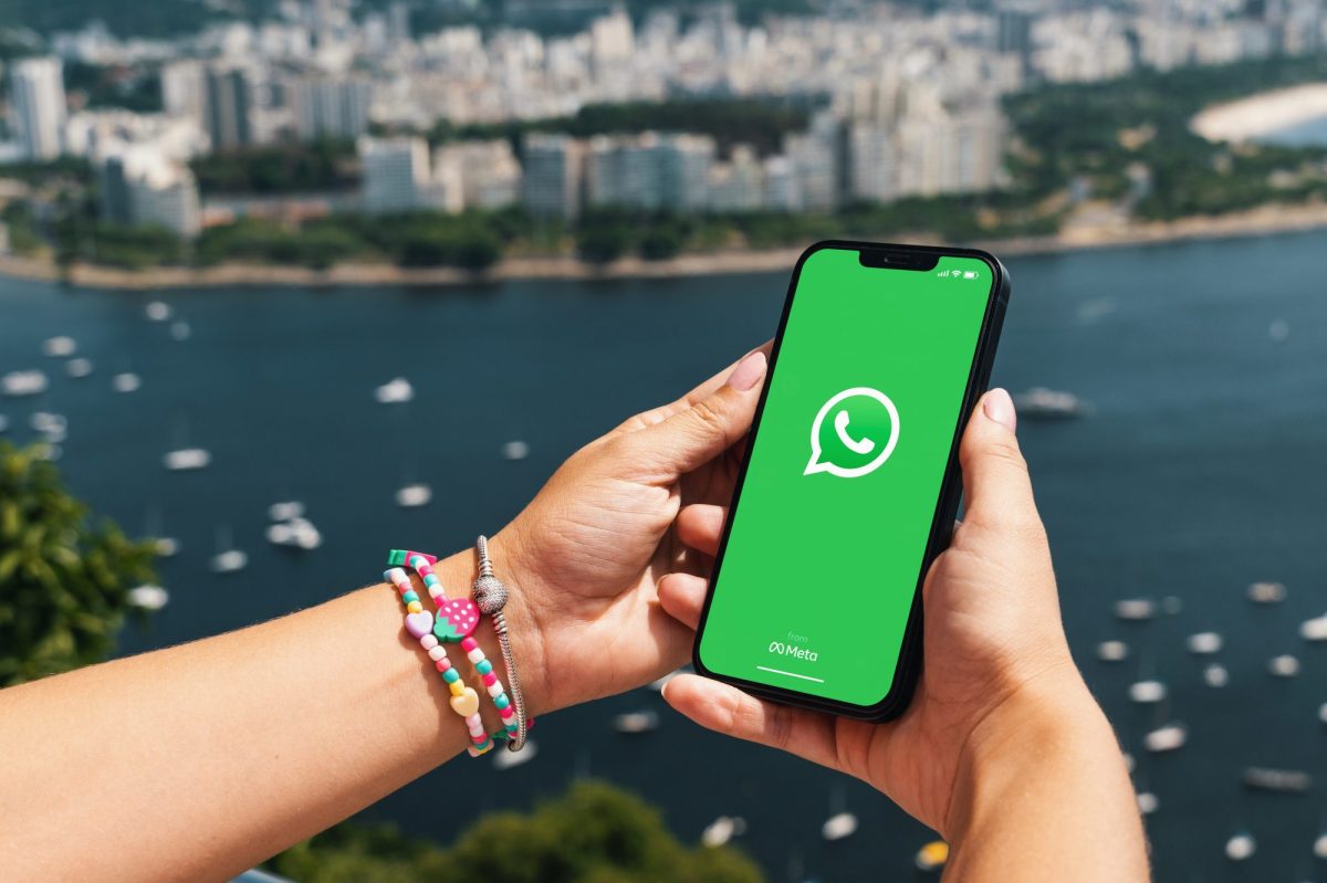 Eine Person hält ein Handy in der Hand, auf dessen Display das WhatsApp-Logo angezeigt wird. Im Hintergrund ist das Meer und die Skyline einer Stad zu sehen.