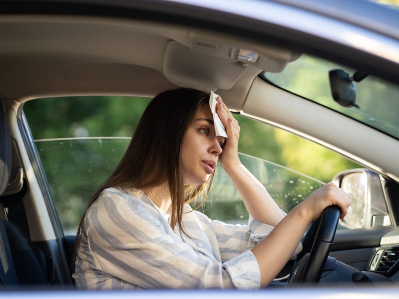 Eine Frau sitzt im Auto und hält sich ein Tuch an den Kopf und stützt sich auf das Lenkrad. Das Autofenster ist leicht geöffnet.