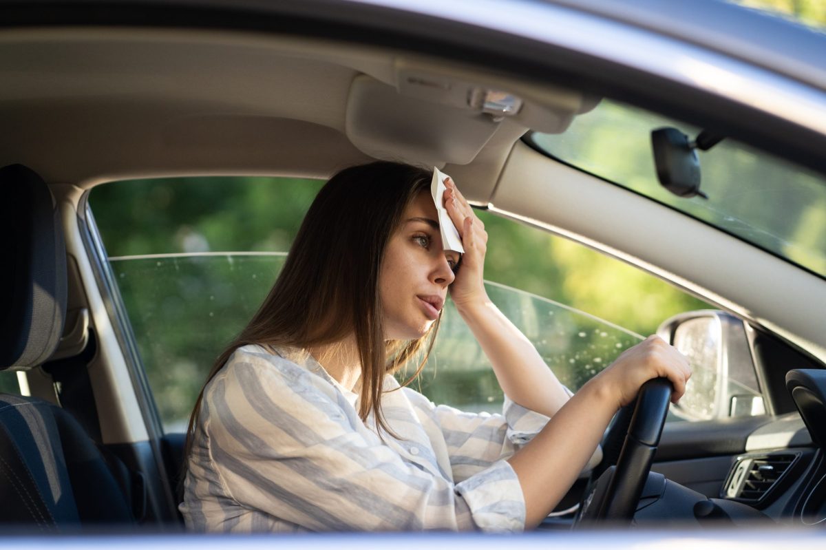 Eine Frau sitzt im Auto und hält sich ein Tuch an den Kopf und stützt sich auf das Lenkrad. Das Autofenster ist leicht geöffnet.