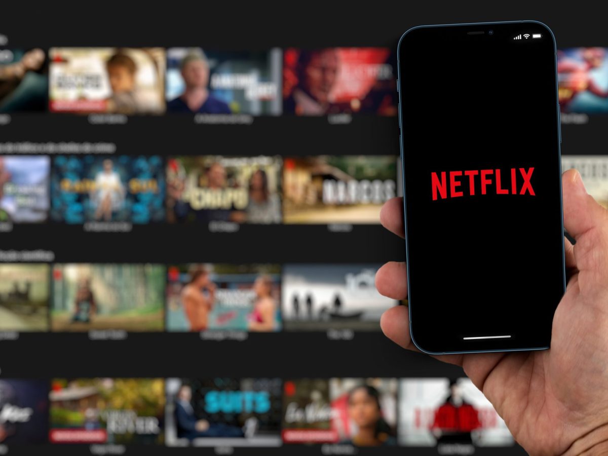 Handy zeigt Netflix-Logo vor einem Fernseher als Hintergrund.