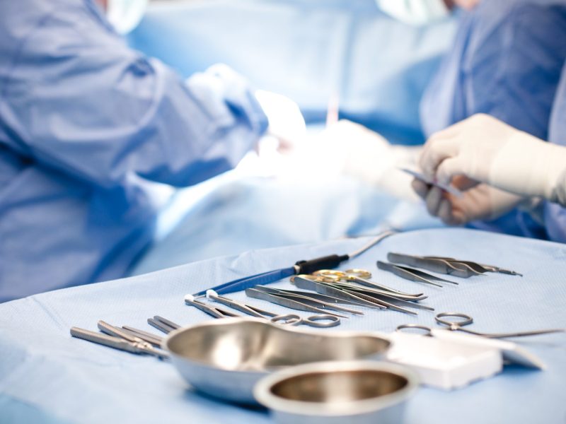 Auf einem Tisch liegen Operations-Instrumente. Im Hintergrund operieren Chirurgen einen Patienten.