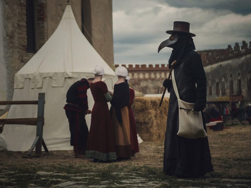 Ein Pest läuft an einer mittelalterlicher Burg entlang. Im Hintergrund sind drei Personen vor einem weißen Zelt in mittelalterlicher Kleidung zu sehen.