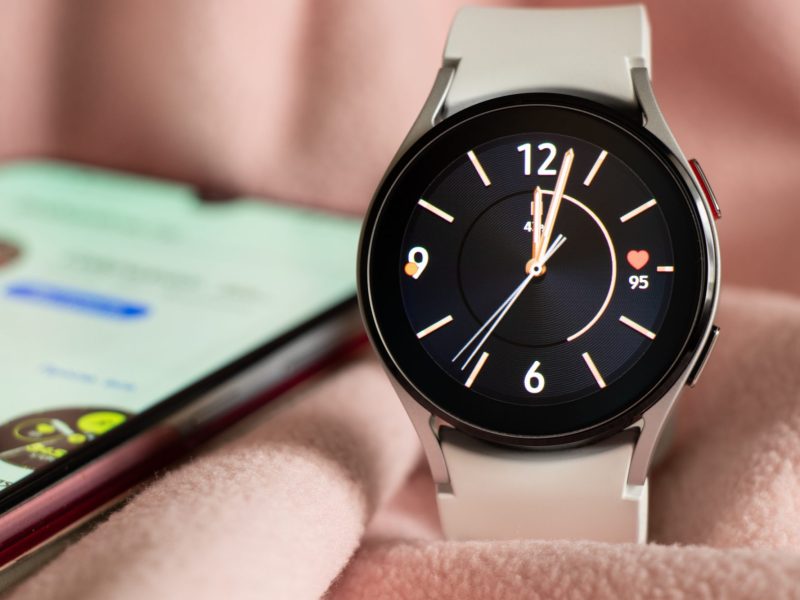 Samsung Galaxy Watch liegt auf einem rosafarbenen Untergrund. Im Hintergrund liegt ein Smartphone.