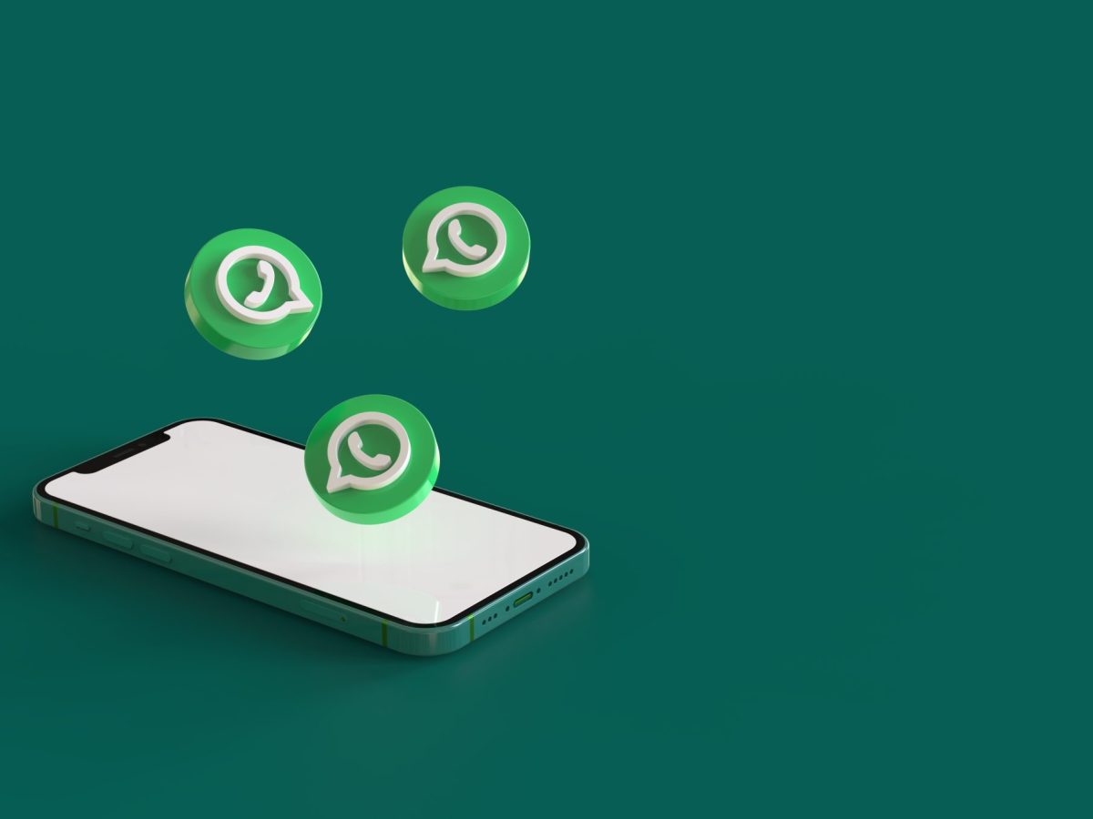 Ein Handy liegt auf einem grünen Untergrund. Darüber schweben drei WhatsApp-Logos.