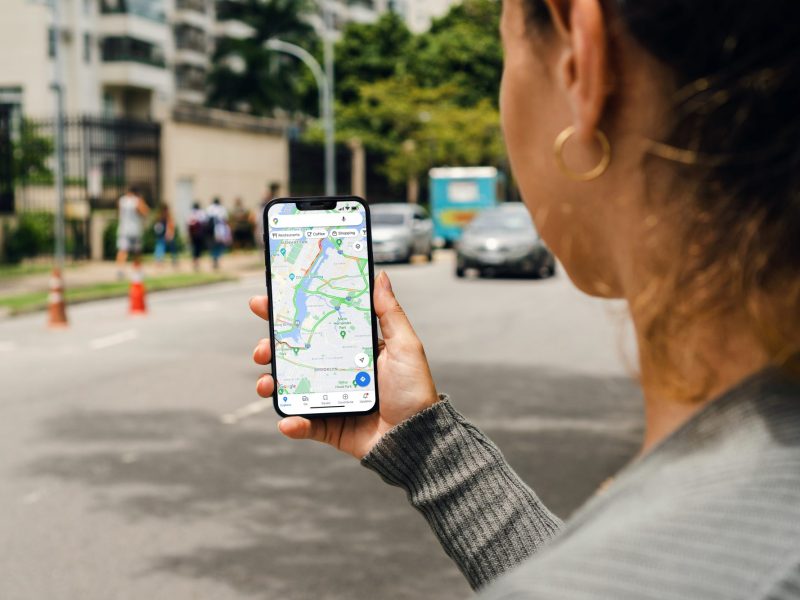 Person hält Handy in der Hand, auf dem Google Maps geöffnet ist. Im Hintergrund sind Straßen und Häuser zu sehen.