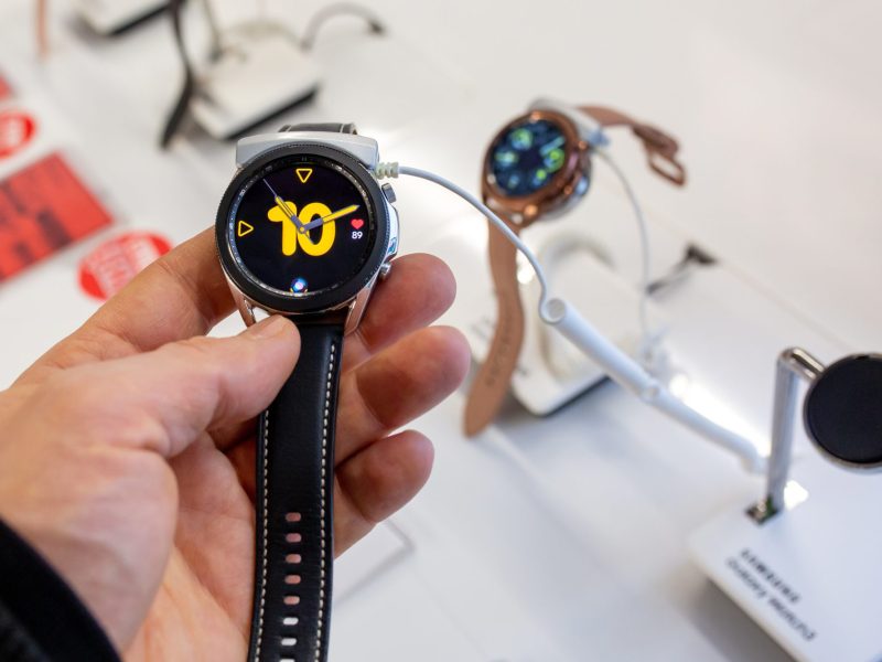 Eine Person hält eine Samsung Galaxy Watch in der Hand. Im Hintergrund sind in einer Auslage weitere Samsung Galaxy Watch-Modelle zu sehen.