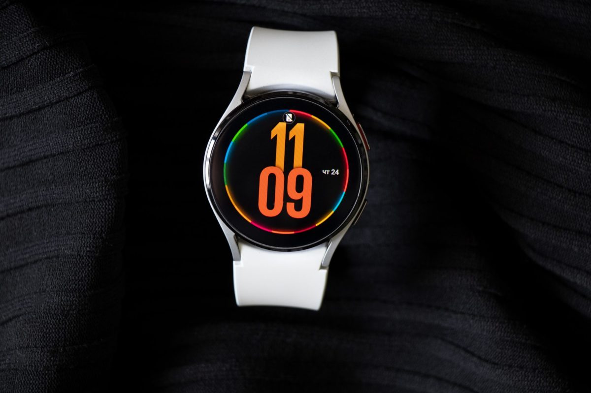 Samsung Galaxy Watch4 liegt auf einem dunklem Untergrund. Auf dem Ziffernblatt wird die Uhrzeit in oranger Schrift angezeigt.