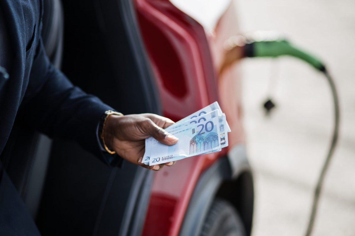 Person hält Geldscheine in der Hand. Im Hintergrund wird gerade ein Elektroauto geladen.