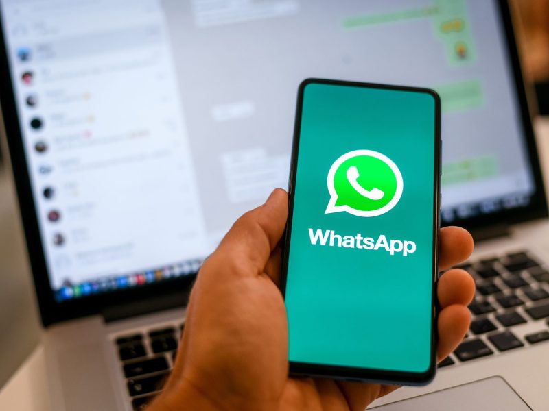 Person hält Handy in der Hand. Auf dem Bildschirm ist das WhatsApp-Logo zu sehen, im Hintergrund steht ein Laptop, auf dem WhatsApp geöffnet ist.