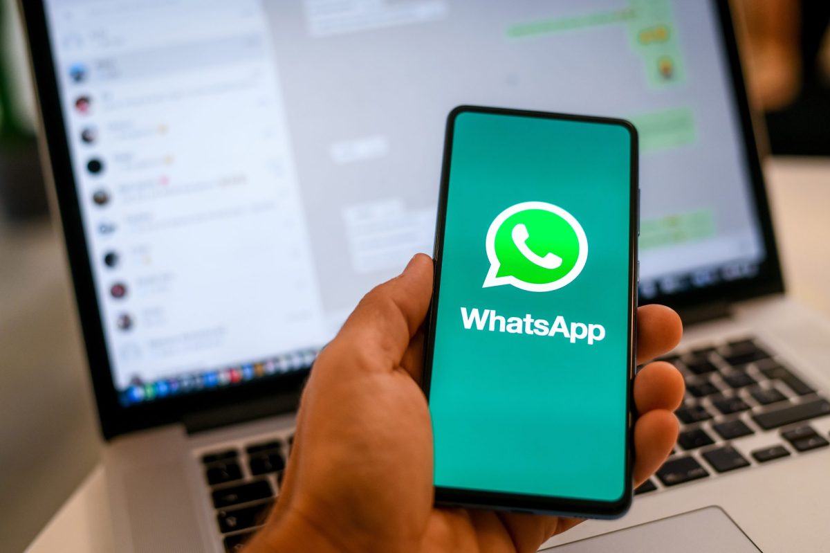 Person hält Handy in der Hand. Auf dem Bildschirm ist das WhatsApp-Logo zu sehen, im Hintergrund steht ein Laptop, auf dem WhatsApp geöffnet ist.