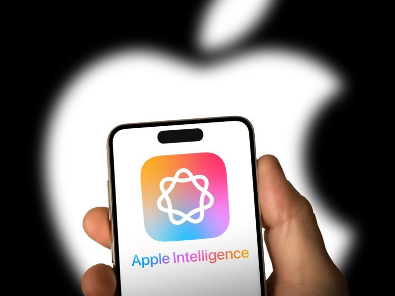 Person hält ein Handy. Auf dem Display wird das Apple Intelligence-Logo angezeigt. Im Hintergrund ist das Apple-Logo zu sehen.