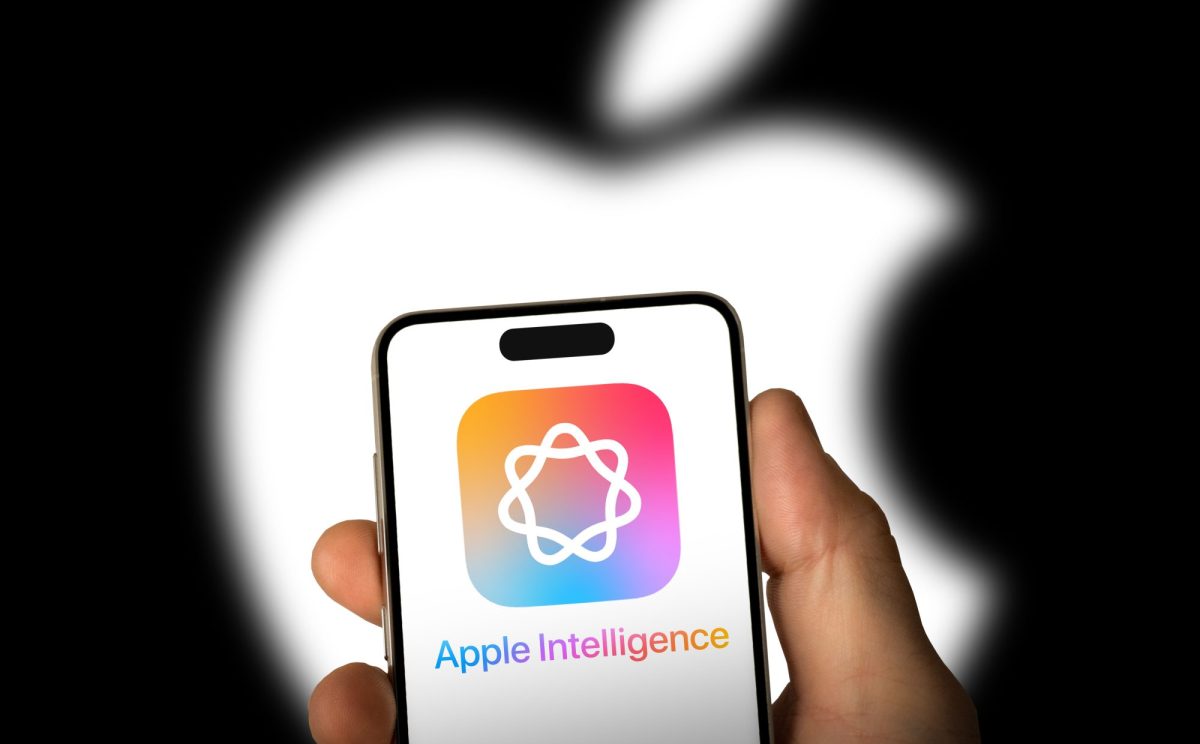 Person hält ein Handy. Auf dem Display wird das Apple Intelligence-Logo angezeigt. Im Hintergrund ist das Apple-Logo zu sehen.