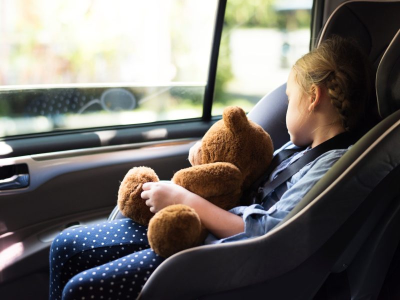 Mädchen sitzt in einem Auto-Kindersitz und hält einen Teddy in der Hand, während sie aus dem Fenster guckt.