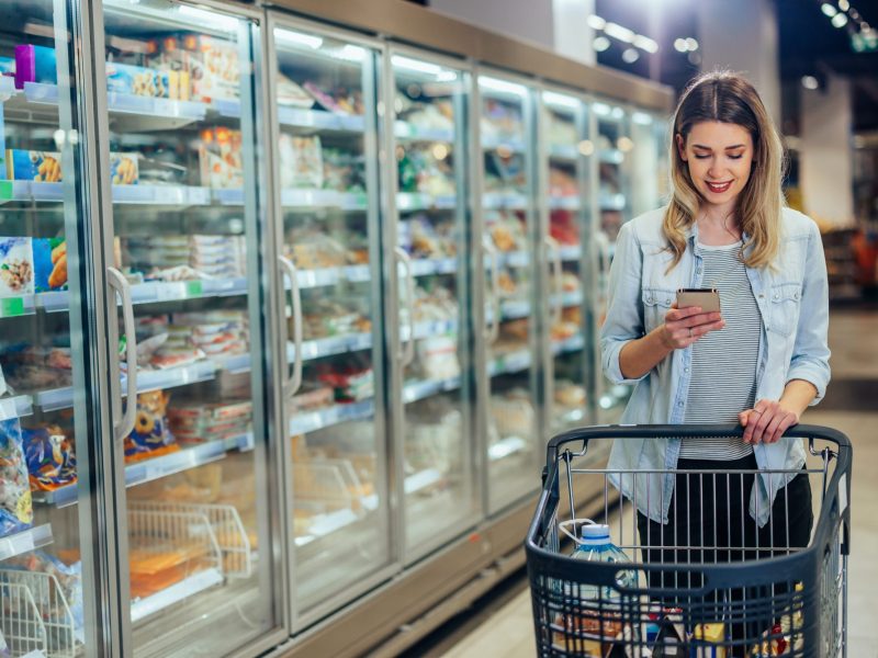 Frau steht im Supermarkt und blickt auf ihr Handy.