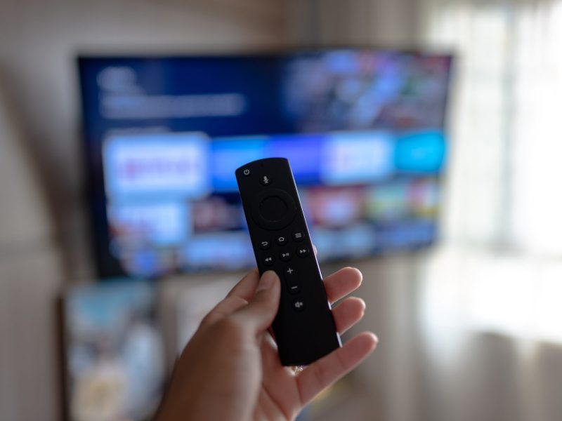 Person hÃ¤lt die Fernbedienung des Amazon Fire TV Sticks auf einen angeschalteten Fernseher gerichtet.