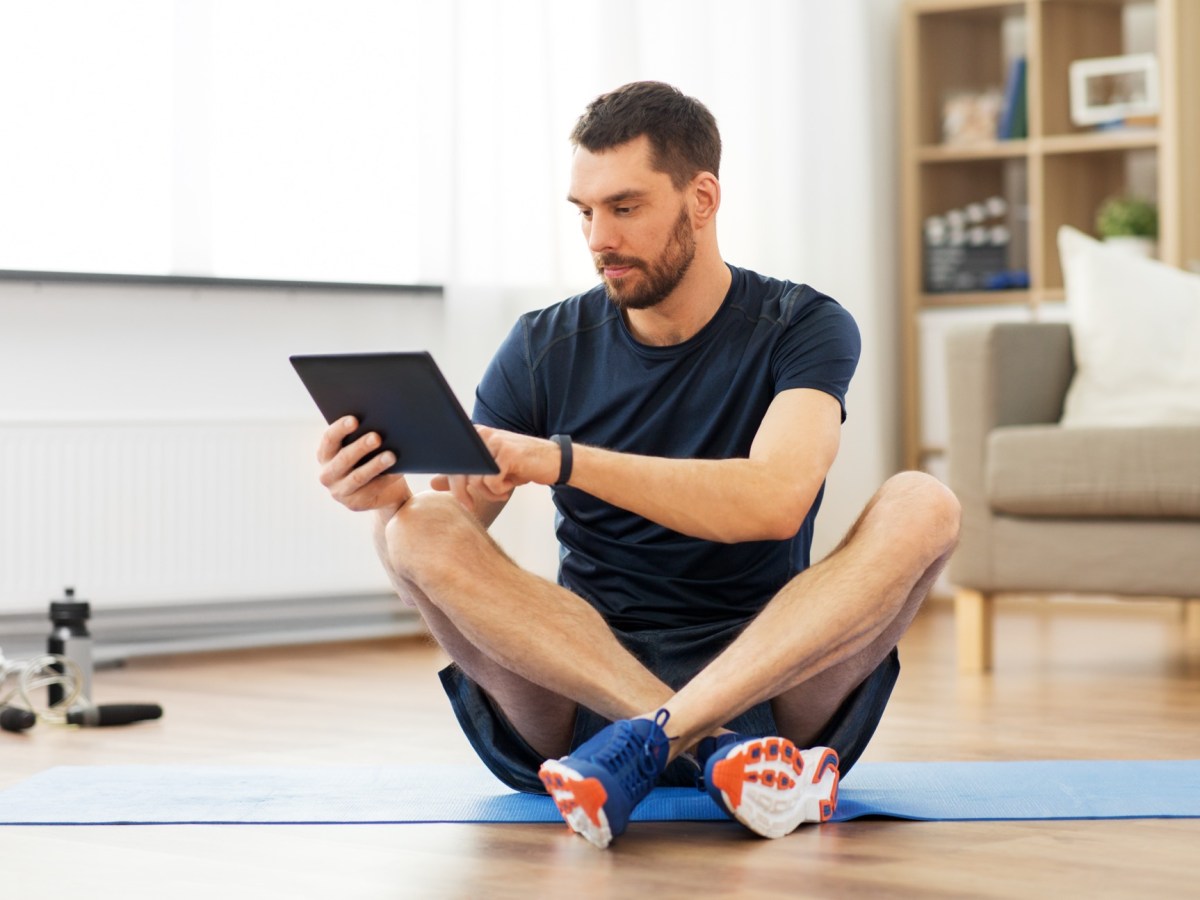 Mann schaut auf sein Tablet, wÃ¤hrend er auf einer Fitness-Matte sitzt.