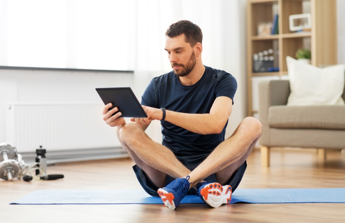 Mann schaut auf sein Tablet, wÃ¤hrend er auf einer Fitness-Matte sitzt.