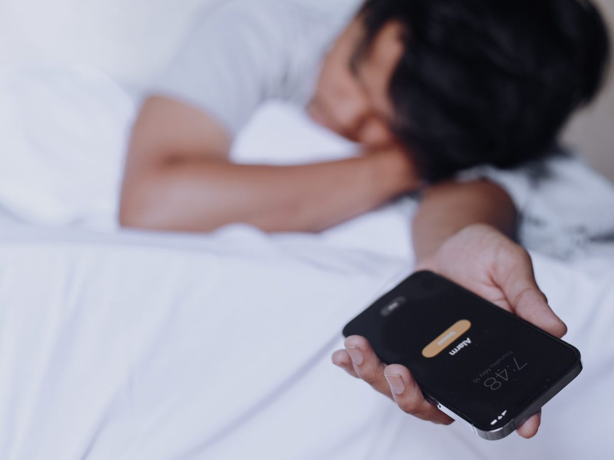 Person liegt im Bett und hÃ¤lt ein Handy in der Hand, auf dem ein Wecker klingelt.