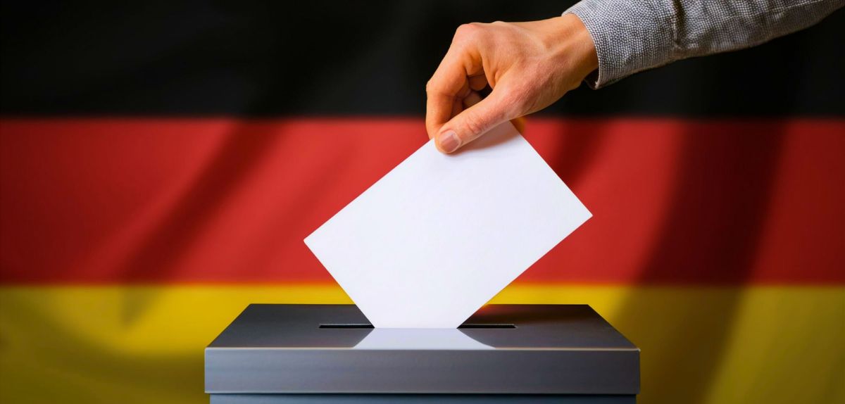 Person wirft Zettel in die Wahlurne (vor einer Deutschlandfahne)