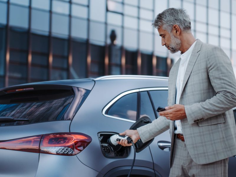 Eine Person im Anzug hÃ¤lt ein Handy in der Hand und lÃ¤dt ein Elektroauto. Im Hintergrund ist ein modernes GebÃ¤ude mit Glasfront zu sehen.