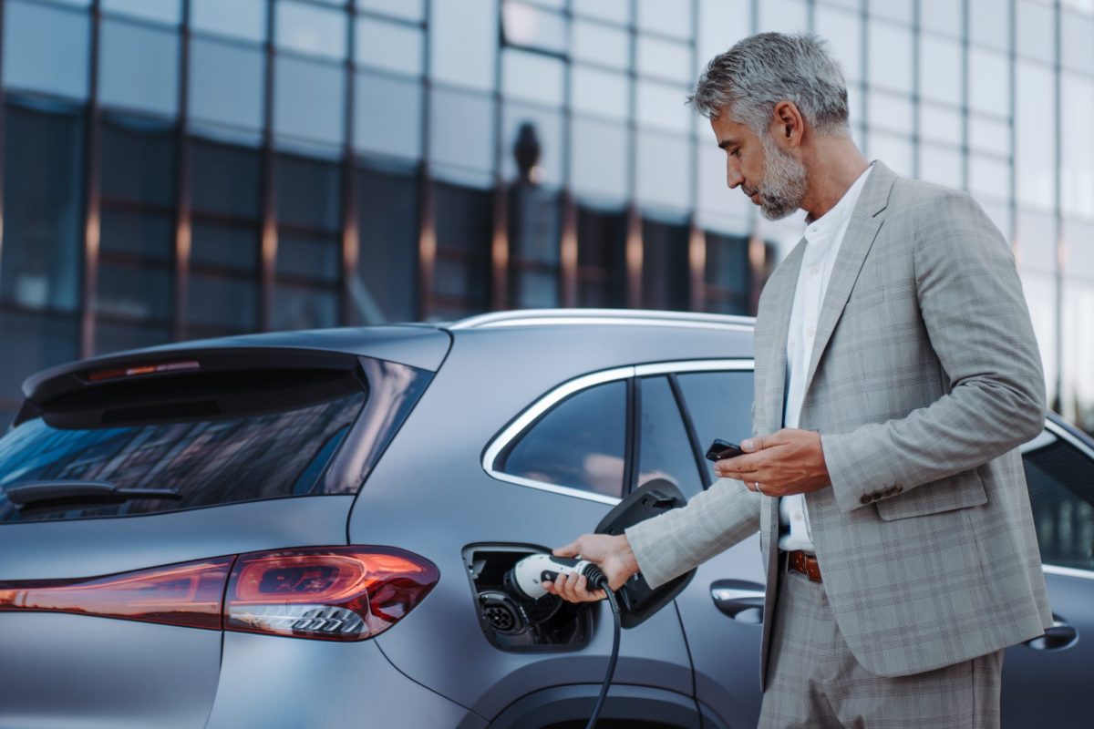 Eine Person im Anzug hÃ¤lt ein Handy in der Hand und lÃ¤dt ein Elektroauto. Im Hintergrund ist ein modernes GebÃ¤ude mit Glasfront zu sehen.