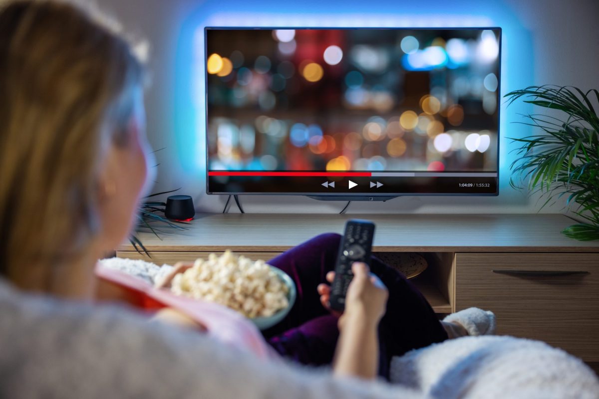 Frau sitzt mit Fernbedienung in der Hand und Popcorn auf dem SchoÃŸ vor einem Fernseher.