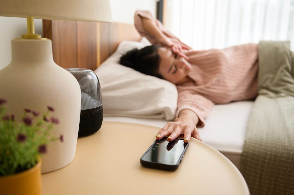 Eine Person liegt im Bett und greift zum Handy, das auf einem Tisch neben dem Bett liegt.